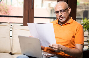 Man wearing an orange shirt looking at papers in front of his laptop.