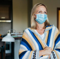 Blonde woman wearing mask and sweater standing in living room.