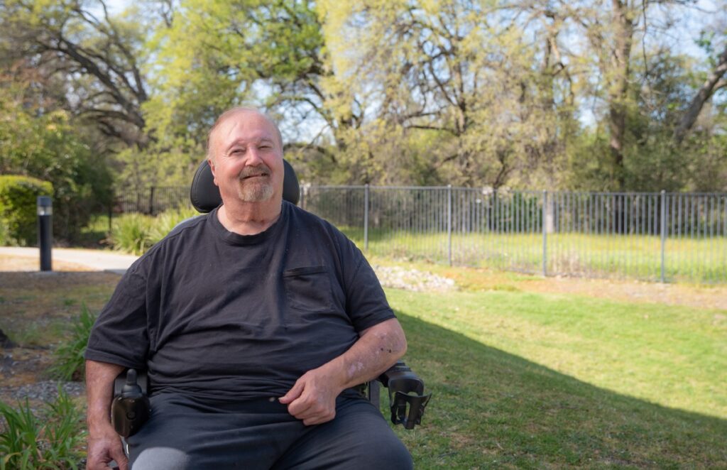 Man in motorized wheelchair outside in a park, smiling.
