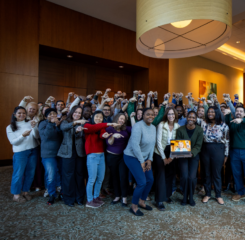 Nearly 50 Leadership Academy Fellows pose for group photo, smiling and pointing to laptop with woman joining by Zoom