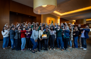 Nearly 50 Leadership Academy Fellows pose for group photo, smiling and pointing to laptop with woman joining by Zoom